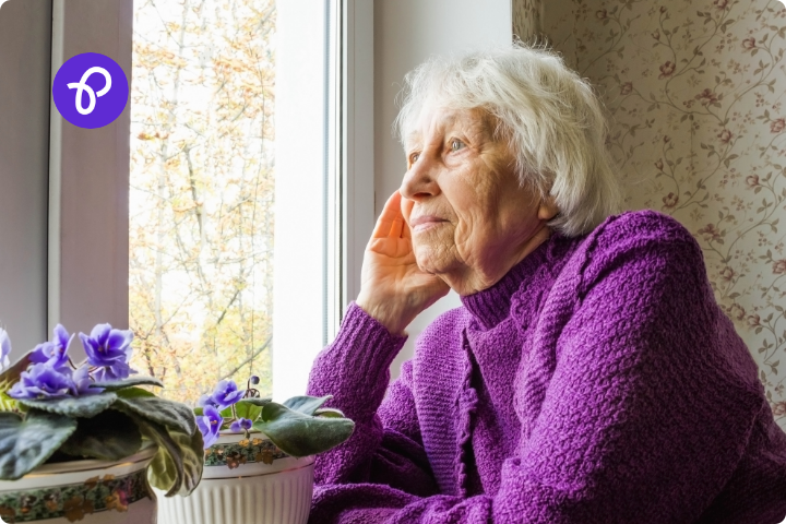 An older white woman with white hair and a purple jumper looks sadly out of a window.