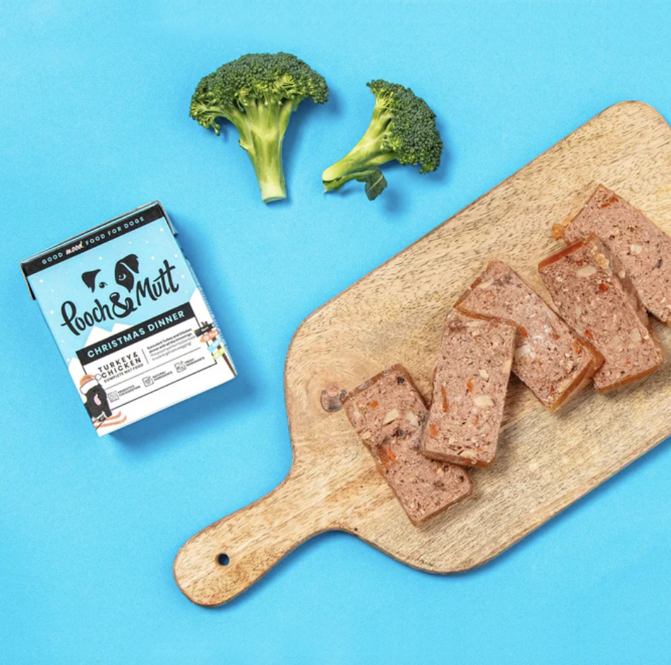 A bright blue background with a chopping board with sliced dog food, a pooch and mutt pouch and some broccoli 