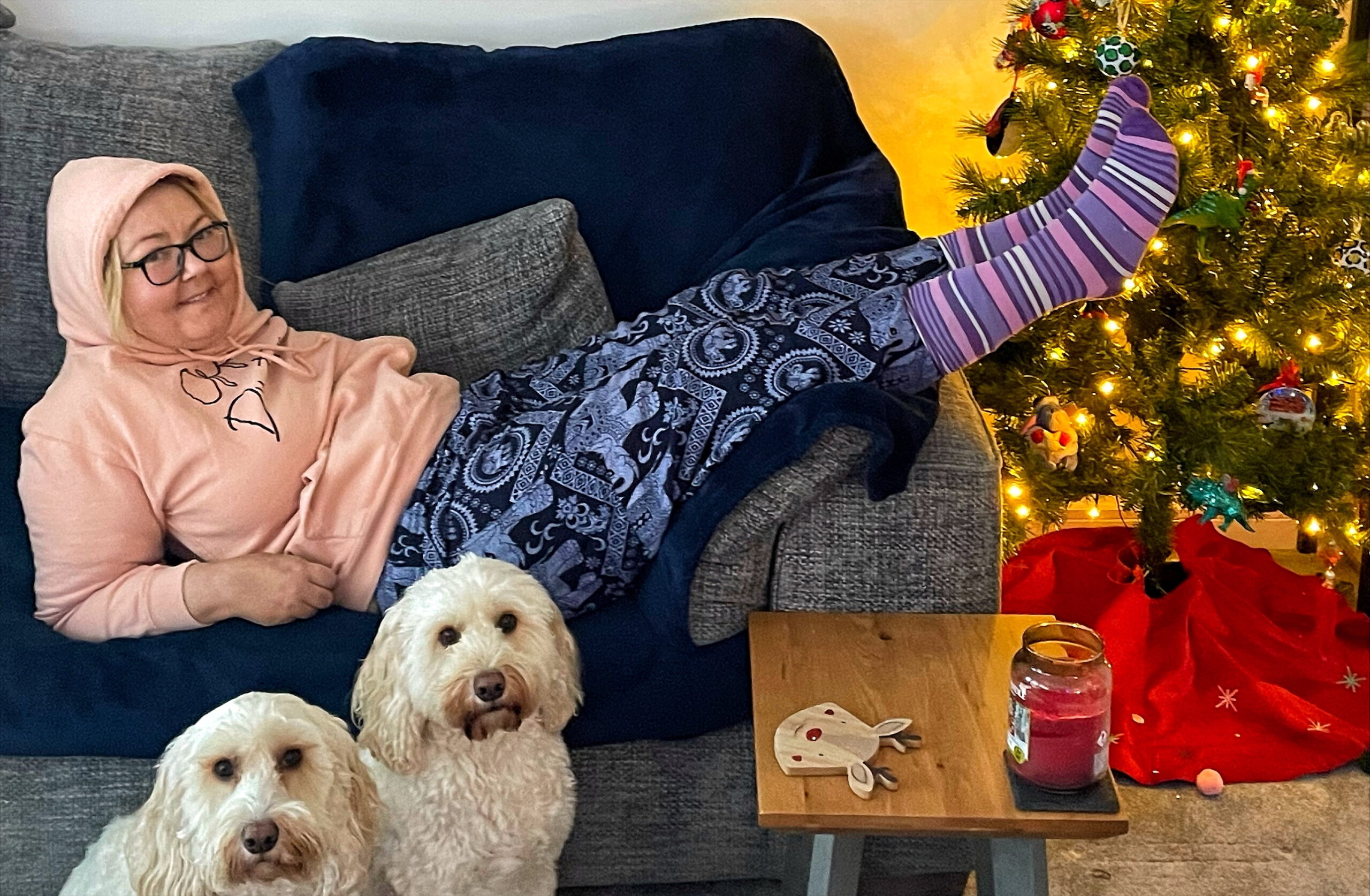 Christy is a white woman wearing a peach hoodie and purple socks, she is laid on the sofa by a christmas tree with two white fluffy dogs sat by her side