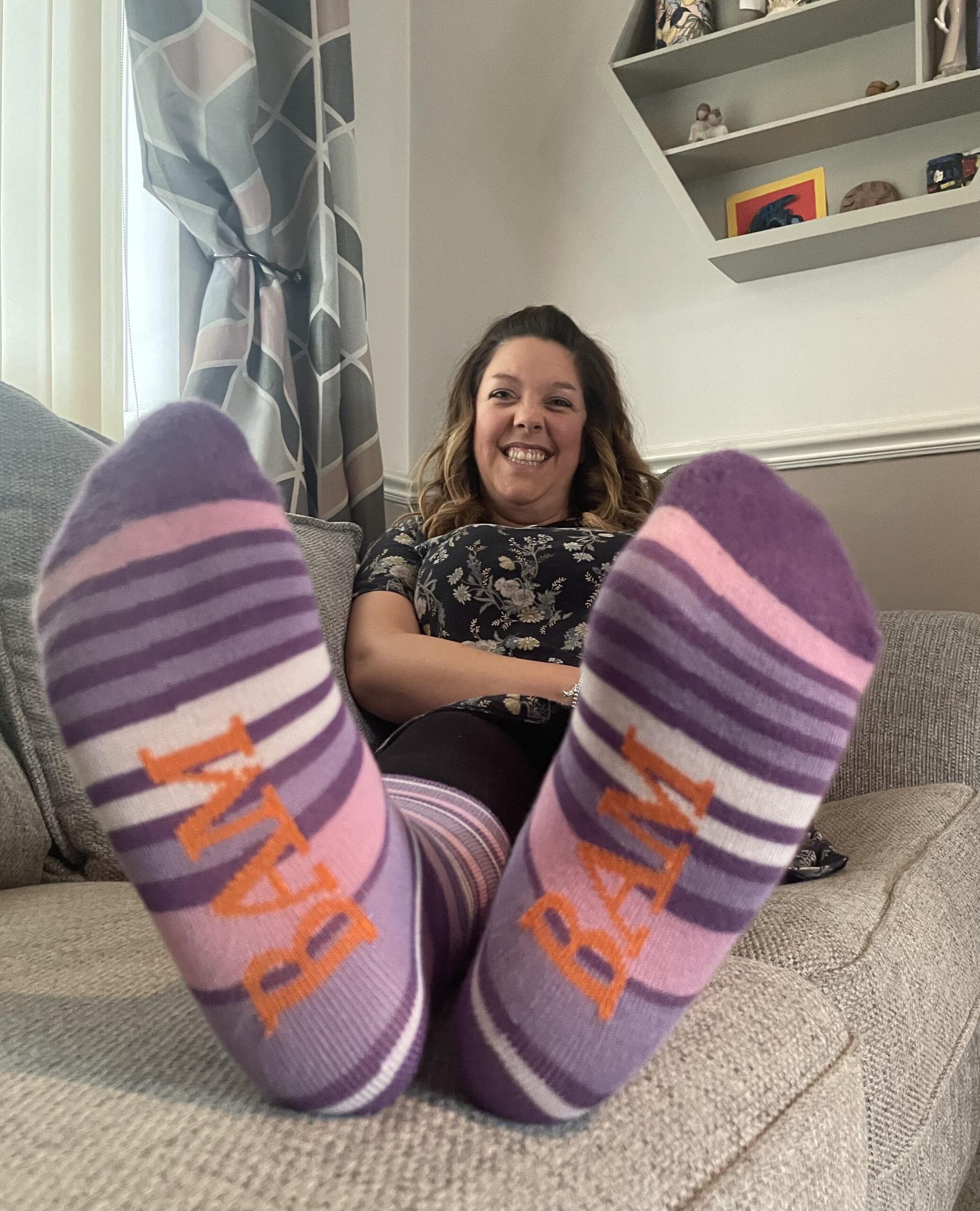 A white woman with shoulder length hair smiles to camera, her feet are in the forefront of the image and she is celebrating Purple Sock Day for the International Day of Disabled People celebrating Purple Sock Day for the International Day of Disabled People 