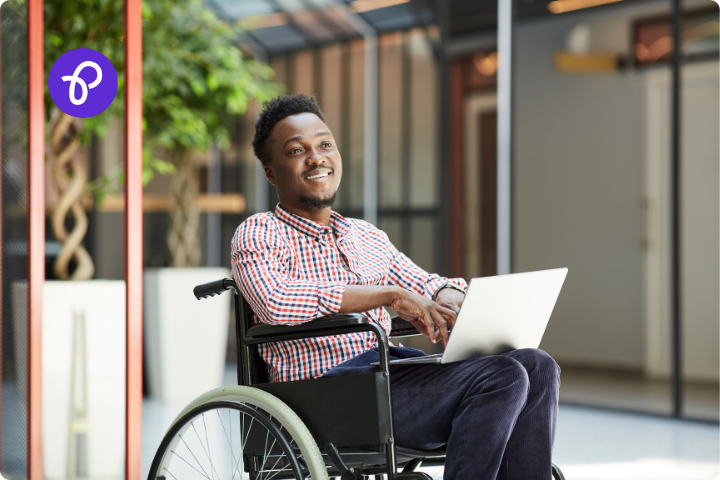 A black man with short black hair wearing a checked shirt, he is a wheelchair user and has a laptop on his knee, he is outside an office space and is smiling