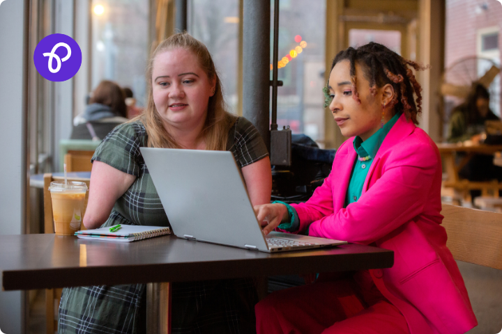 A white woman with a limb difference is sat at a table with a black woman, they are both looking at a laptop.