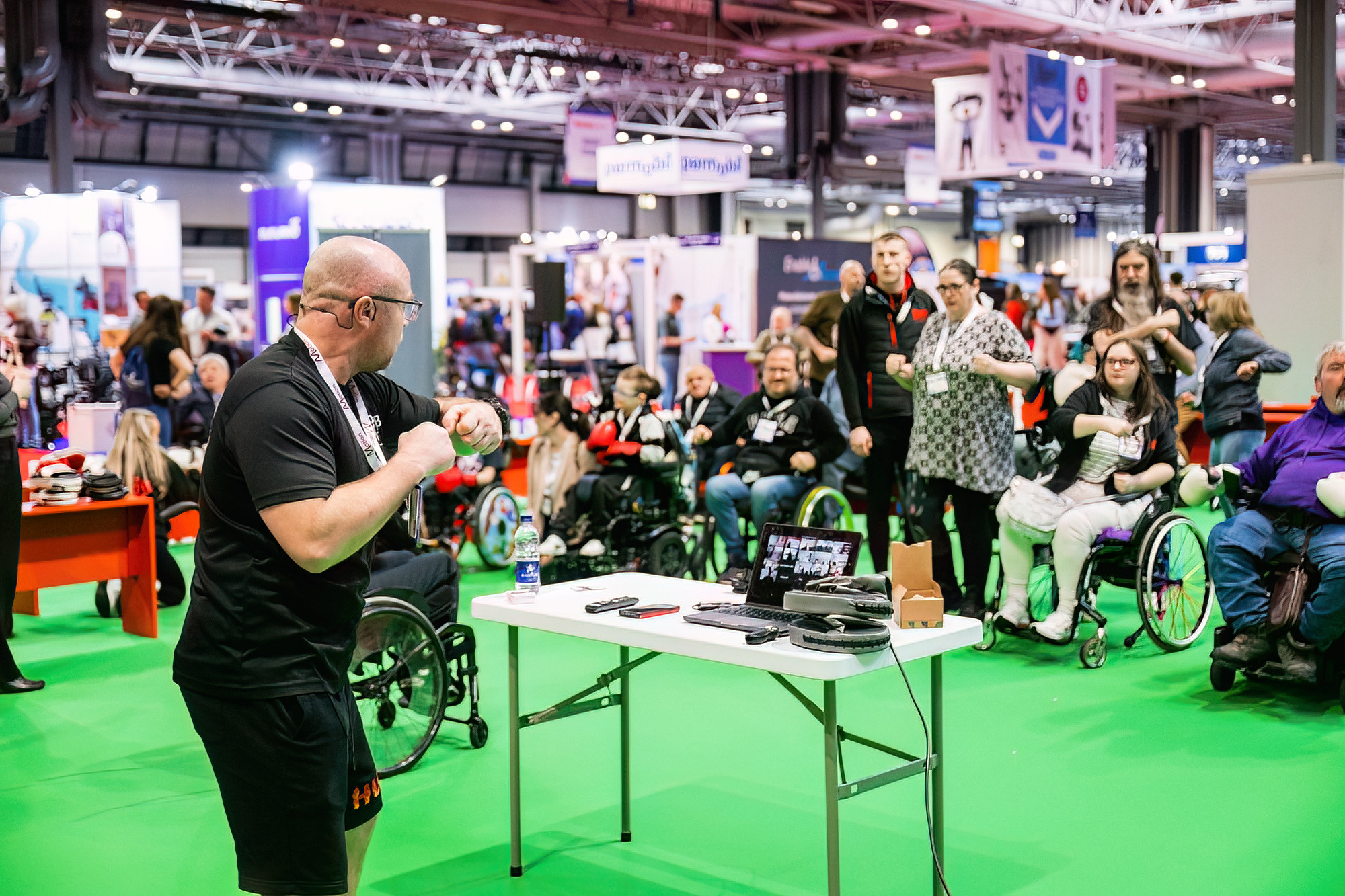 A bald man dressed in black is in front of a group of people at a Naidex event, he appears to be demonstrating some boxing techniques, in the background there are a range of people, some wheelchair users who are copying his pose.