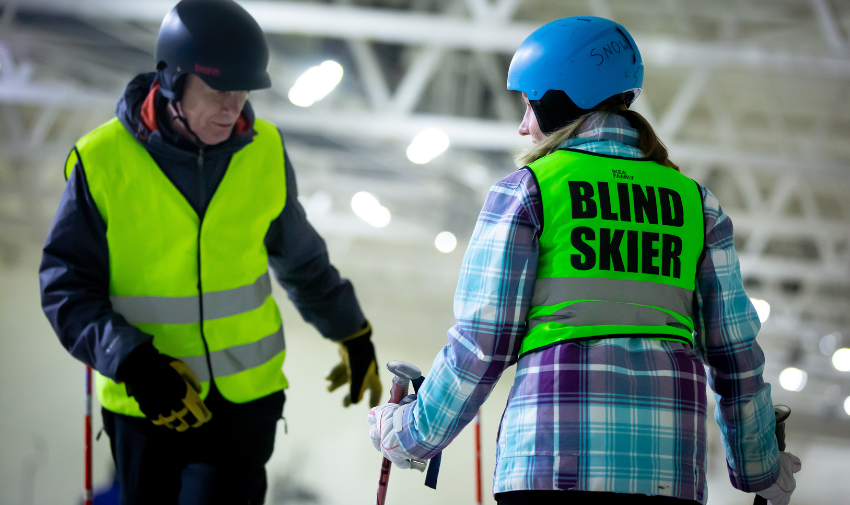 Two people in warm clothes, helmets and high vis waistcoats, the man is facing towards the camera snd the other person has their back to camera and on the back of their waistcoat it says Blind Skier, the background is blurred but it looks like they are at an indoor ski slope