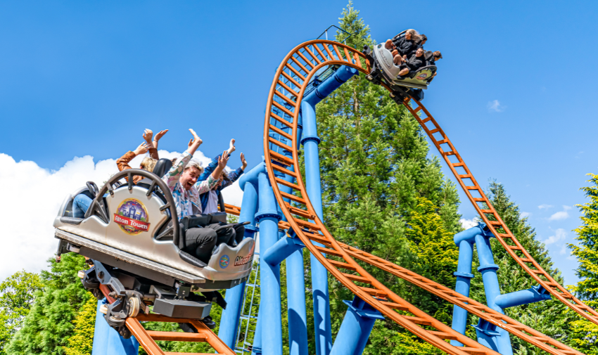 An Alton Towers rollercoaster, there are orange rails on blue frame and people in small carriage sat back to back, they have their arms in the air and look excited and happy.