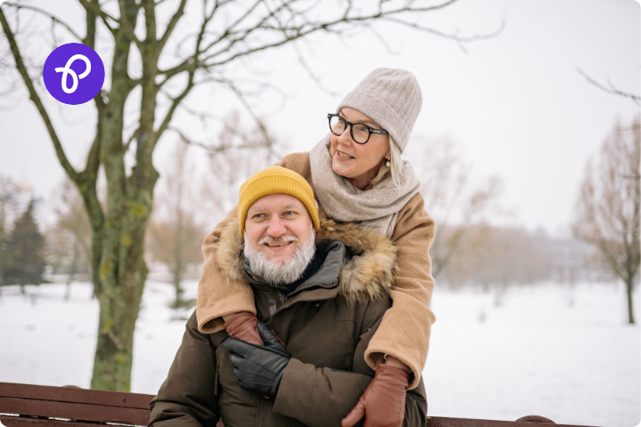 A male female white couple are outdoors in the snow, the man has a white beard and a yellow hat and is sat on a bench, the woman wears glasses and a cream hat and scarf and stand behind him with her arms around his shoulders