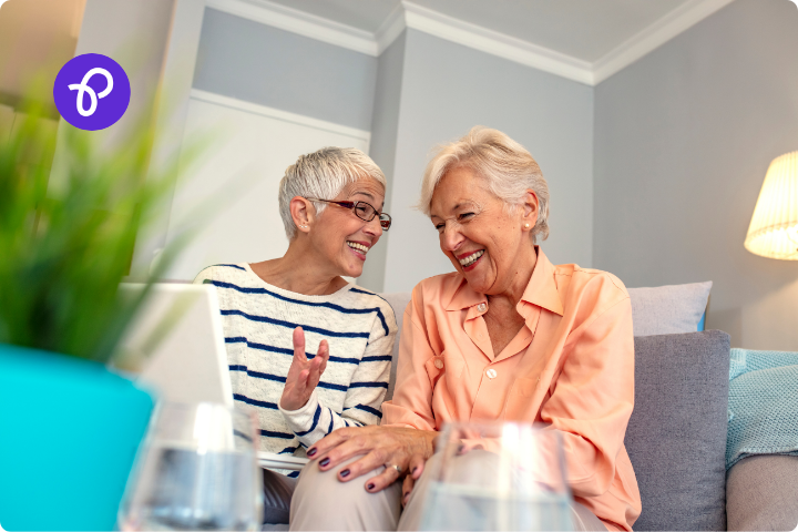 An older female white couple are sat on a sofa together laughing, the woman on the left has white cropped hair, glasses and a white and black striped top, the woman on the right has short silver hair and wears a peach blouse.