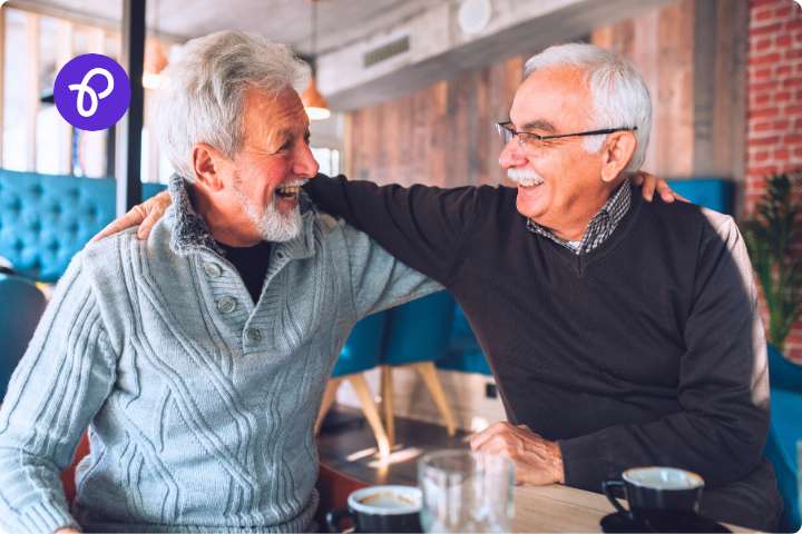 An older male couple are sat in a restaurant or cafe, they have their arms around each others shoulders and are smiling at each other. The man on the left has a short grey beard and wears a pale blue ribbed knitted jumper and the man on the right has a white mustache and white hair, wears glasses and a dark jumper over a checked shirt collar.