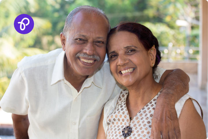 An asian older couple stand together smiling, the man has a mustache and a wears a short sleeved white shirt and the woman has dark hair tied back, in the background are green trees and bushes