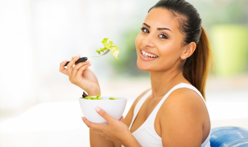 A brown woman with long dark hair pulled back in agony tail is smiling to camera whilst holding a white bowl of salad and a fork with lettuce on it.