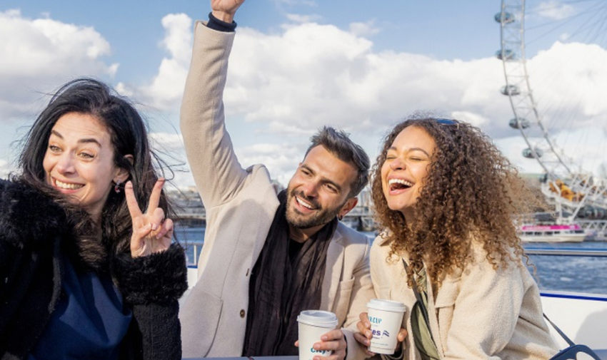 A group of three adults, two women and a man are having fun and posing with peace signs on a city cruise in london, the london eye is behind them