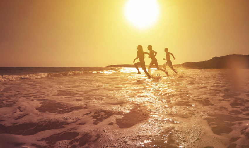 Three silhouettes of people running into the sea, the sun is shining and the who photo has very yellow and orange tones