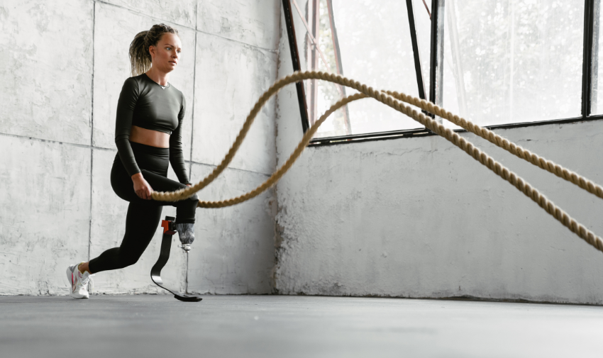 A disabled woman with a lower limb difference is exercising with two ropes in a grey gym, she has a prosthetic leg