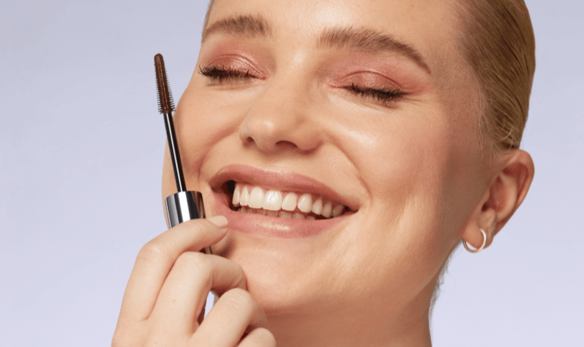 A white woman close up on her face, her eyes are closed and she is wearing natural makeup and holding a mascara wand up to her face