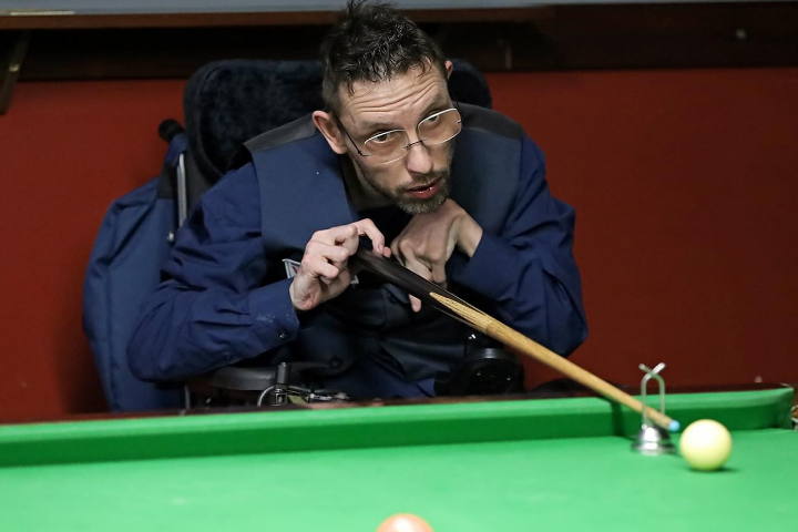 Matt Lester is a white man in his 30s, he has short hair and beard, wears glasses and has cerebral palsy. He is playing snooker at a table with a cue, wearing a waistcoat and shirt and is concentrating