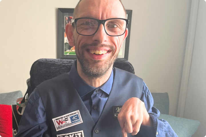 Matt Lester is a white man in his 30s, he has short hair and beard, wears glasses and has cerebral palsy. He is posing to camera wearing a snooker waistcoat and shirt and is smiling