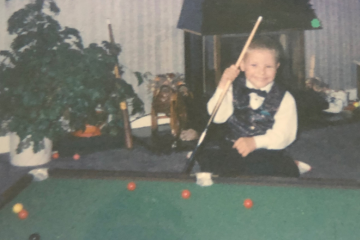 A young boy wearing snooker vest and white shirt and boy tie poses at a small snooker table, the photo is aged and looks like it is from the early 90s.