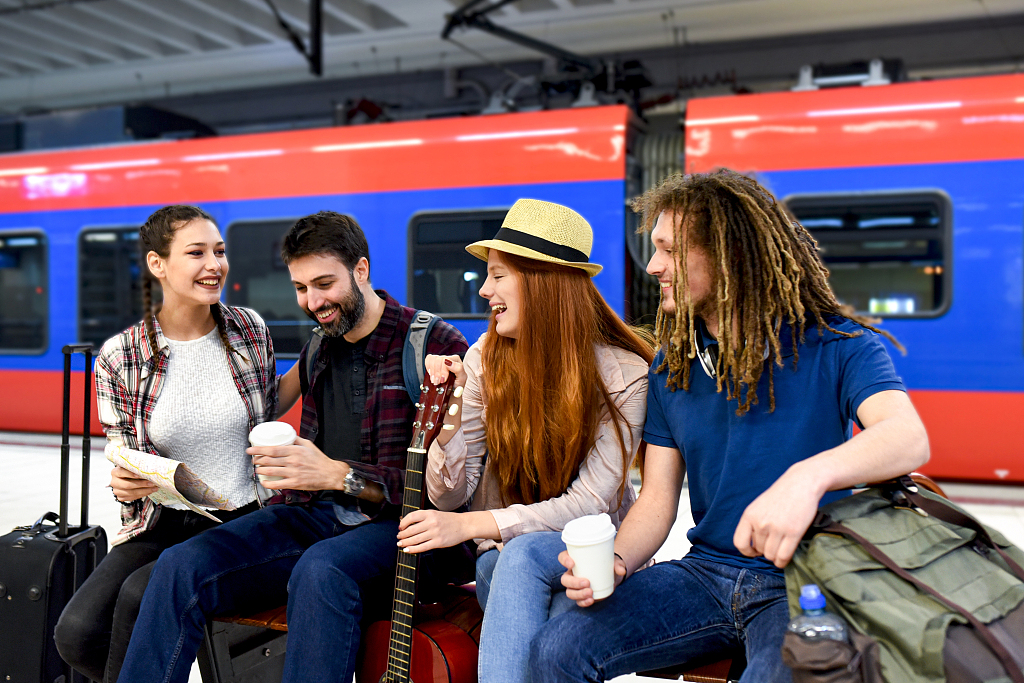 A group of four young adults are sat in a train station with bags and a guitar, they are laughing