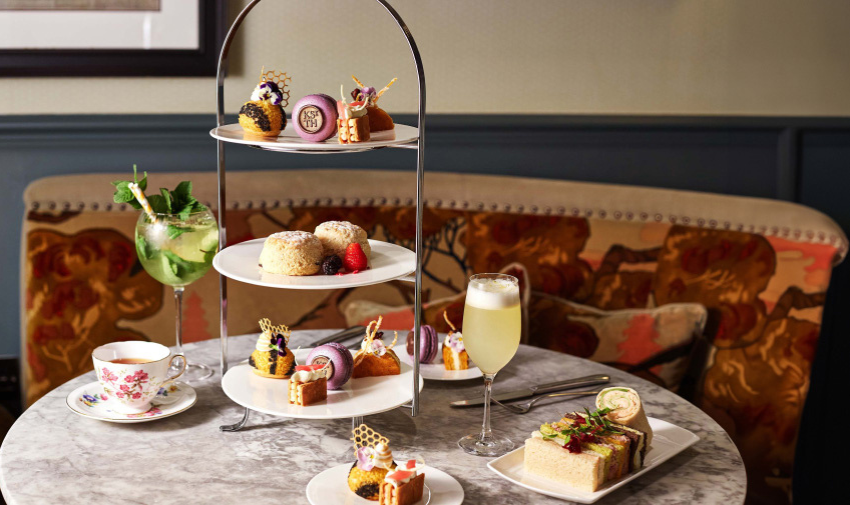 A table is laid out with afternoon tea, sandwiches and cakes are on a tiered tray and there are tea cups and a glass of mimosa on the table too