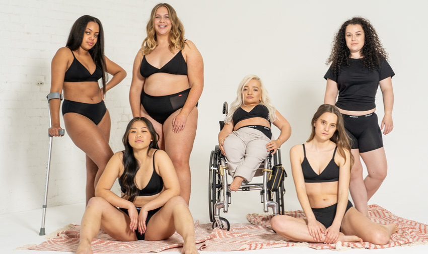 A group of 6 women pose in WUKA underwear, they are different colours, sizes and abilities, one woman is using a crutch and another is a wheelchair user.