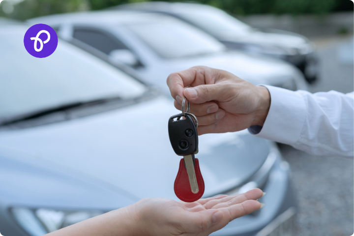 Two hands, one handing over a set of car keys to the other, with a car forecourt in the background