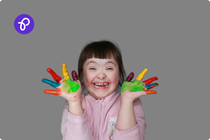 A little girl with special educational needs has brown hair cut into a fringe, she is wearing a pink long sleeved top and have multi colored paint on her hands, she is showing her hands to camera and laughing.