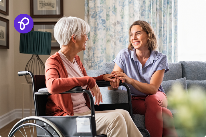 An older woman in a wheelchair is at home with a younger woman who is sat on a safa holding her hand, they are both smiling