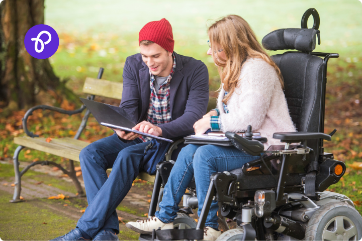 A man and woman are sat together in a park, he is sat on a bench, he wears a suit jacket and a red beanie hat and is holding a folder, she is white and has long fair hair, she is a wheelchair user in a powered chair and is looking at the folder