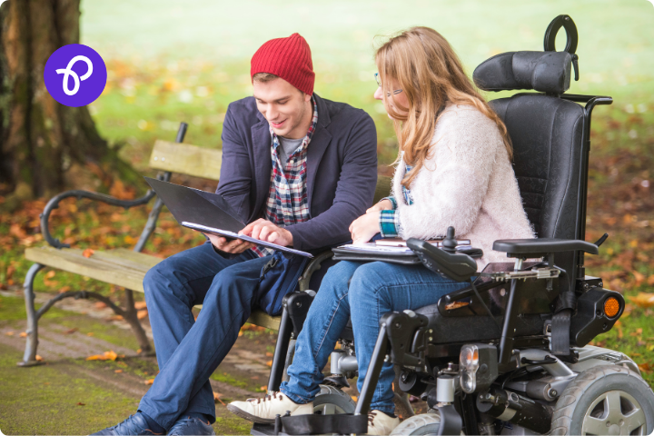 A woman and man sit together in a park looking at a folder of paperwork, the woman has long fair hair and is a wheelchair user, the man sits on a park bench and has a suit jacket and a beanie hat on