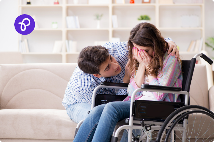 A woman with long dark hair is a wheelchair user, she is in a living room and has her face in her hands looking upset, a man sits on a sofa and has his arms around her in comfort