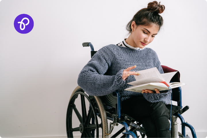 A brown woman is a wheelchair user, she has long dark hair tied up in a bun and a grey jumper, she is looking through a book, the background is plain white