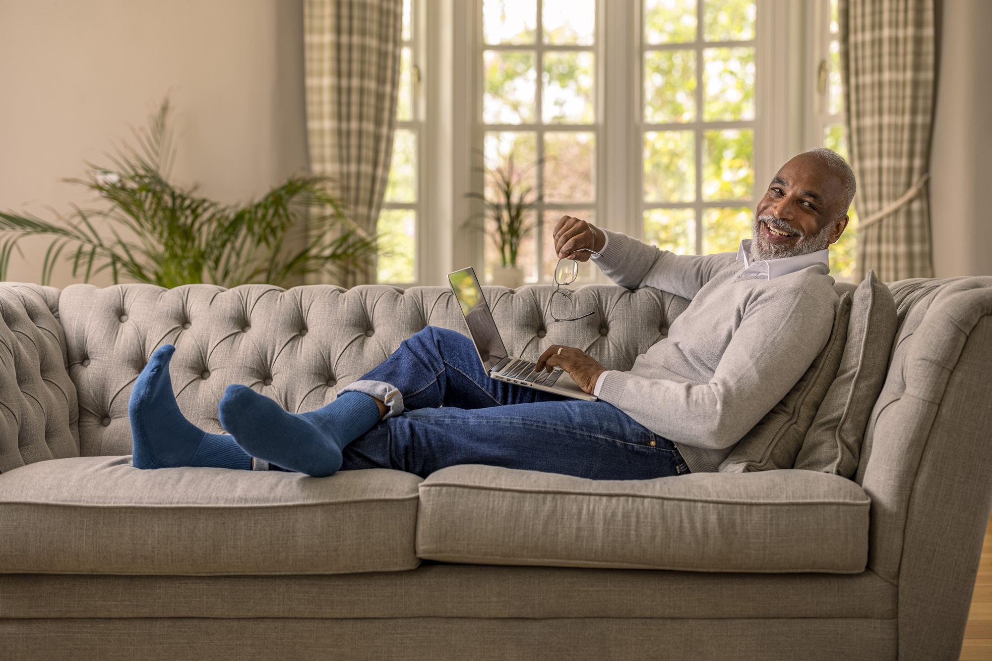 A black man in his 50's is sat back with his legs on a sofa in a living room, he has a laptop on his lap and looks relaxed and happy, he is wearing blue gentle grip socks