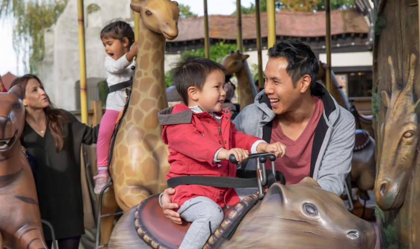 A ride at Chessington world of adventures for small children, there is a little boy in a red coat riding on a capybara, a man is stood next to him keeping him safe, they are asian and look happy and relaxed, behind is a little girl riding a giraffe, a woman stands by her with her hand on her back.