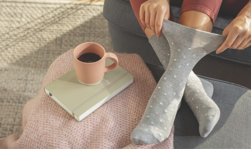 A pair of feet in gentle grip grey socks on a sofa next to a cup and notepad, hands are pulling on one sock