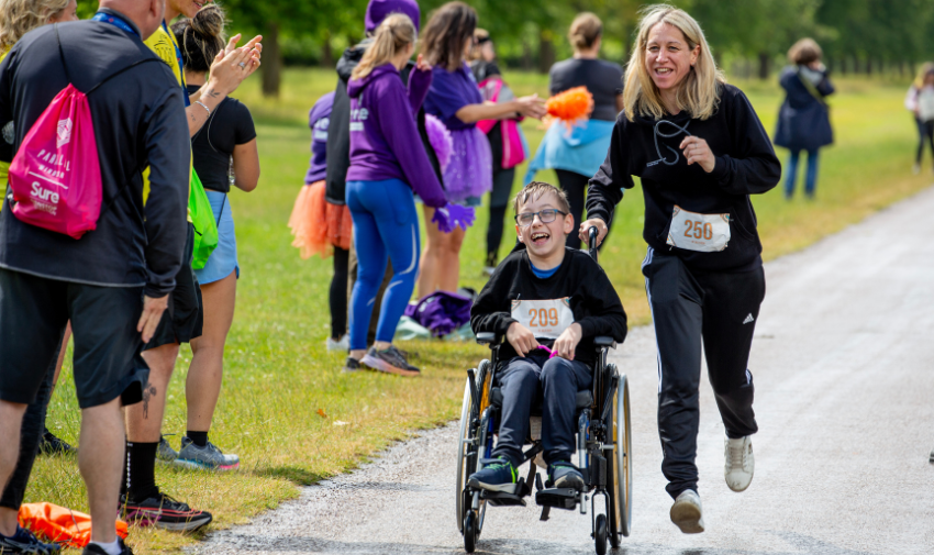 A blonde woman dressed in black with a race number 250 stuck to her top is running and pushing the wheelchair of young boy, he is also dressed in black and has the number 209 on his chest. They are taking part in a fun run at the Parallel Windsor event and are laughing.