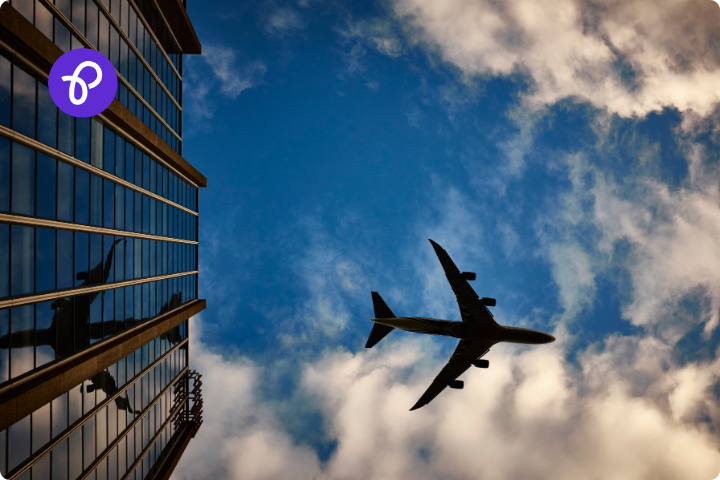 A view upwards to a plane flying through a blue sky near a tall glass building