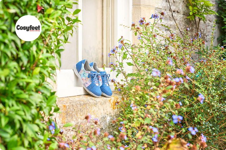 A pair of womens blue and floral trainers are displayed on a windowsill in a garden with lots of greenery and wildflowers around them, there is a Cosyfeet logo in top left hand corner