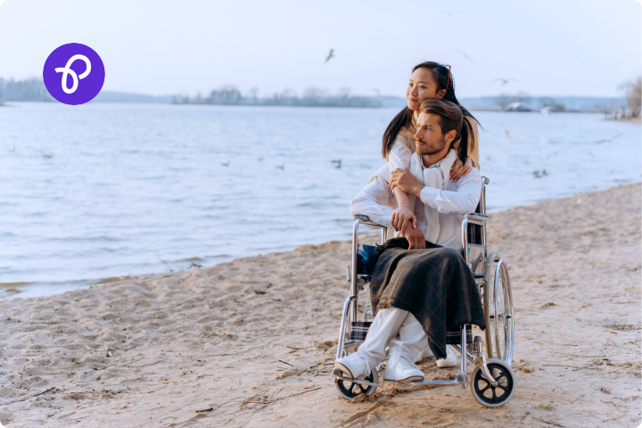 A man and woman are on a beach, the man is a wheelchair user and is wearing white with a dark blanket on his knee, the woman is stood behind him with her arm around him, she has long dark hair