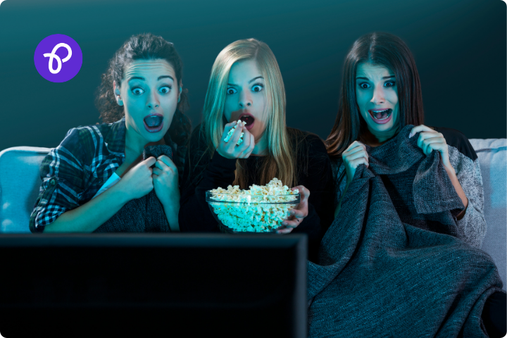 Three women are sat under a blanket on a sofa eating popcorn and watching a movie, they look scared as though they are watching a horror film in a galentines day movie night