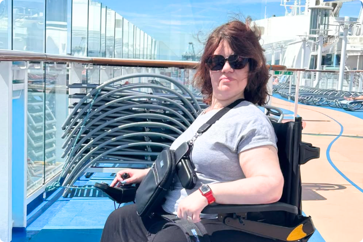 Sara Stacey is a white woman with brown shoulder length hair, she is wearing sunglasses, a grey tshirt and a black bag across her chest, she is a wheelchair user and is on a boat.