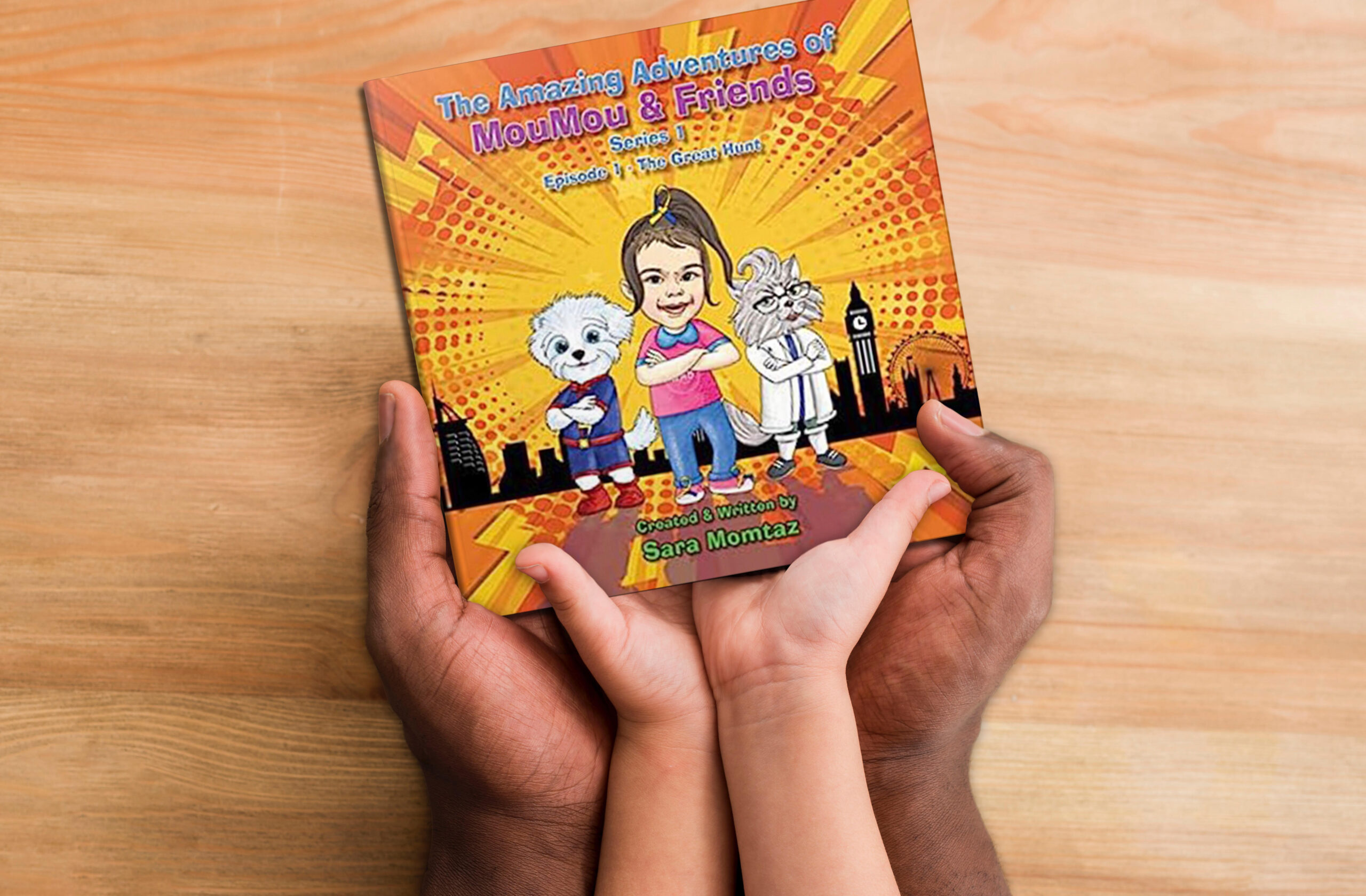 Adult and child hands hold a childrens book called the amazing adventures of moumou and friends, the cover is orange and has a cartoon drawing of a young girl with a ponytail and a dog and cat wearing clothes 