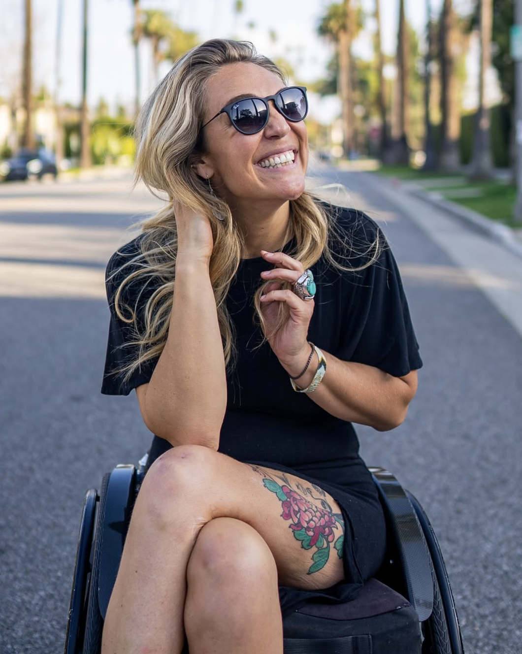 Sophie Morgan is a white woman with long blonde hair, she is a wheelchair user and is wearing sunglasses and a black tshirt, she is on a tree lined road and is smiling