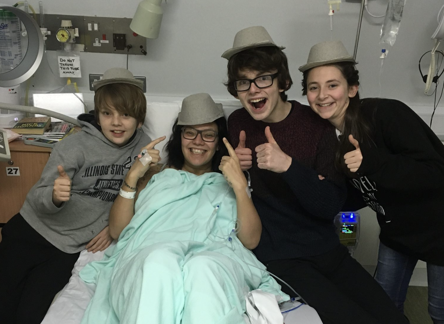 Sam Cleasby is a brown woman, she is in a hospital bed with her three children around her and they all have sick bowls on their heads as hats and are smiling and laughing