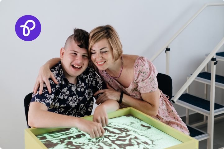 A woman caring for a young adult disabled boy, he is using a sensory table and they are laughing