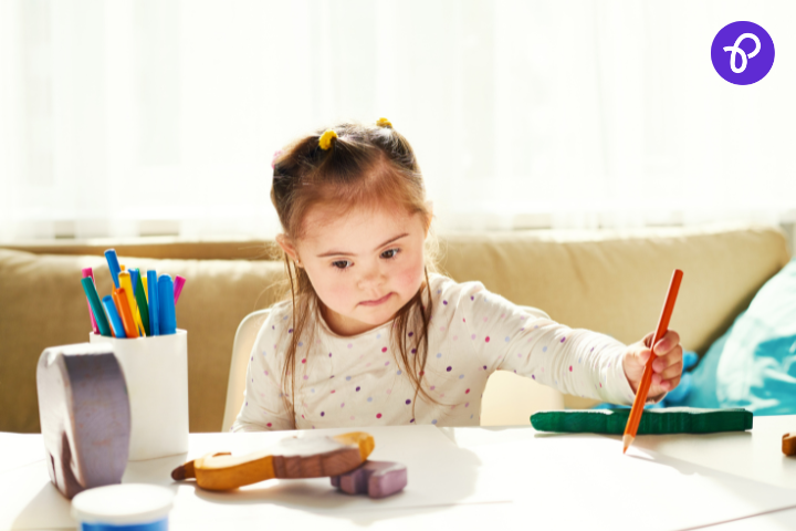 Young girl with Down syndrome sitting at a table in a sunlit room, focused on drawing with an orange pencil, surrounded by colourful pens, modelling clay, and educational toys.