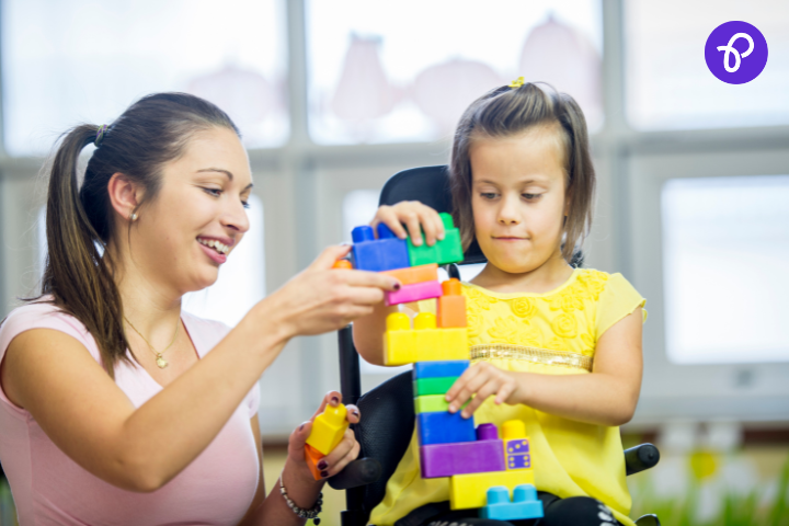 Smiling woman helping a young girl in a wheelchair build a tall tower using colourful plastic blocks in a classroom setting, showing supportive play and development for disabled children.