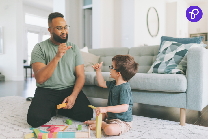 Adult man and young boy wearing glasses sitting on the floor in a bright living room, using sign language while playing with colourful wooden blocks, highlighting inclusive interaction and support for children with disabilities in the UK.