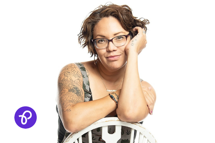 Sam Cleasby is a woman in her 40s of indian and british descent, she has short curly brown hair and glasses and is sat backwards on a chair wearing dungarees