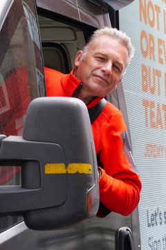 Chris Packham leaning out of a van window wearing a bright red jacket, smiling at the camera, with part of a campaign message visible on the digital billboard beside him.