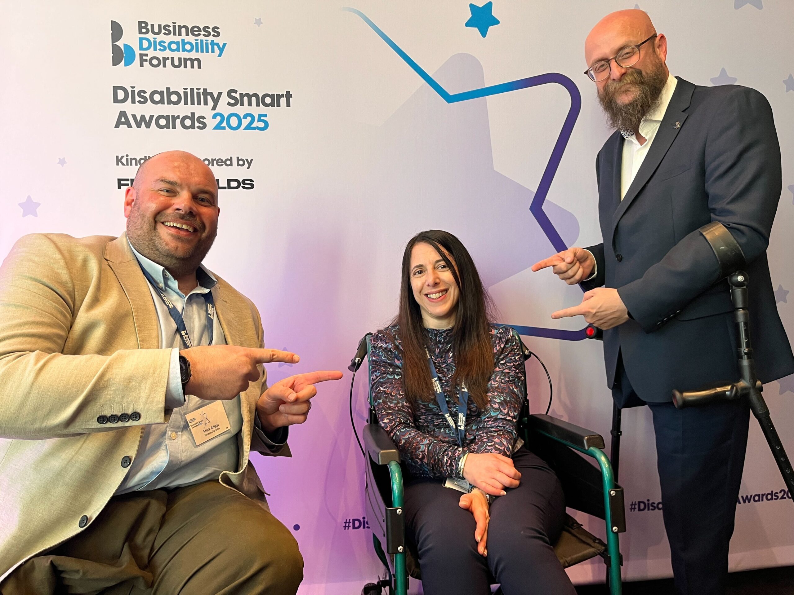 Picture showing Georgina smiling in her wheelchair at the Disability Smart Awards, following her win as the Disability Smart Leader of the Year 2025. To her right and left are two men in suits, smiling and pointing at Georgina.